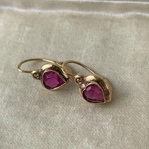 May include: A pair of gold earrings featuring heart-shaped, faceted pink-red stones. Each earring has a curved wire hook and a small, round decorative accent at the top of the heart setting, showcasing a polished metal finish and vibrant gemstone.