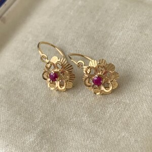 May include: A pair of gold earrings featuring a flower-shaped design with a central round red gemstone. Each earring has a textured, scalloped petal edge and a curved wire hook closure, presented inside a cream-colored velvet-lined jewelry box.