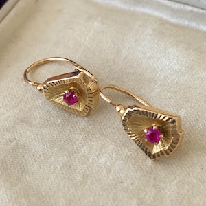 May include: A pair of gold earrings featuring a textured, fan-shaped design with a single round, vibrant pink gemstone set in the center of each piece. The earrings are equipped with secure lever-back closures for attachment to the earlobe.