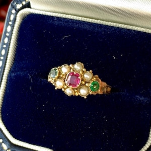 May include: A gold ring featuring a central square-cut red gemstone flanked by two smaller green gemstones, surrounded by a cluster of small white pearls. The ring is presented inside a dark blue velvet-lined jewelry box with white trim.