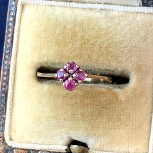 May include: A gold ring featuring a four-stone cluster of round, deep pink rubies arranged in a flower-like pattern. The ring is displayed inside a vintage-style, square-shaped jewellery box lined with beige velvet fabric.