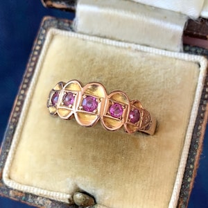 May include: A gold ring featuring five round, vibrant pink gemstones set in a row. The band has a scalloped, decorative edge design surrounding each stone, with engraved detailing visible on the side of the metal band near the setting.