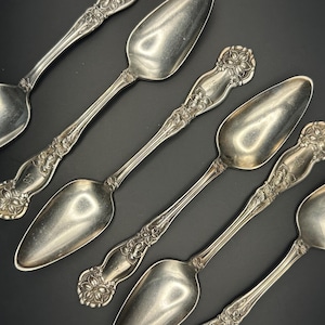 May include: An overhead shot displays six antique silver spoons. Each spoon features an ornate floral design on the handle and a wide, oval bowl. The spoons are arranged diagonally against a dark background, showcasing their reflective surfaces.