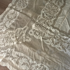 May include: A sheer, cream-colored fabric with a floral and leaf pattern. The design is repeated along the edges, creating a scalloped border. The fabric is slightly translucent, allowing the background to show through.