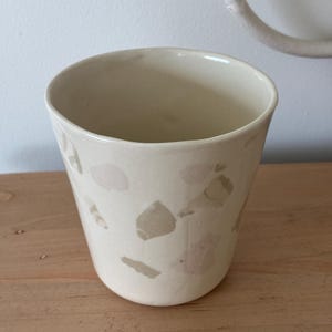 May include: Cream-colored ceramic tumbler with a speckled design in shades of pink and tan. The cup has a rounded base and a slightly flared rim. The tumbler is likely used for drinking beverages.
