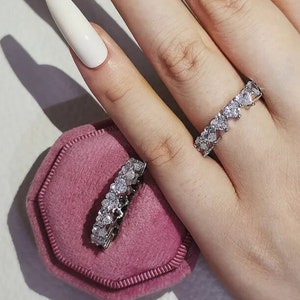 May include: Two silver rings featuring heart-shaped stones. One ring is worn on a finger, and the other rests on a pink velvet ring box. The rings are adorned with multiple clear stones, creating a sparkling effect.