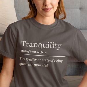 May include: A charcoal grey t-shirt featuring the word "Tranquility" in white, with its phonetic spelling and definition. The definition states "The quality or state of being quiet and peaceful."