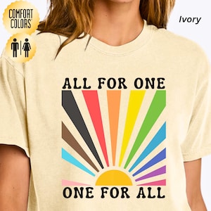 Può includere: T-shirt color avorio con la scritta nera "ALL FOR ONE ONE FOR ALL". Il design presenta un sole colorato con raggi arcobaleno. La maglietta ha un'etichetta Comfort Colors.