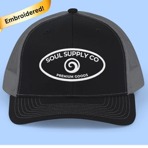 May include: Black and gray trucker hat with the embroidered text "SOUL SUPPLY CO PREMIUM GOODS" in white. The hat has a curved brim and a mesh back. The word "Embroidered!" is on a gold banner.