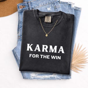May include: Black t-shirt with the text "KARMA FOR THE WIN" in white, paired with blue jeans and a gold necklace. The shirt is made of a soft material. A straw hat and a dried palm leaf are also visible.