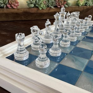 May include: A clear glass chess set with a blue and white checkered board. The chess pieces are transparent and detailed, including pawns, rooks, knights, bishops, a queen, and a king. The board is framed in white.