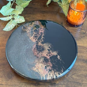 May include: A round, black resin tray with a glossy finish. The tray features a unique design with silver, gold, and pink accents, creating an abstract, flowing pattern. The tray is a decorative item, suitable for serving or display.