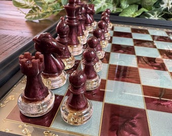 May include: A chess set with burgundy and clear pieces on a checkered board. The pieces are arranged on the board, with the rooks, knights, bishops, and pawns visible. The board has a black frame and a mix of burgundy and light blue squares.