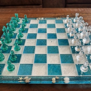 May include: A decorative chess set with a blue and white checkered board. The chess pieces are clear and teal-colored. The board is decorated with seashells and starfish along the border. The set is displayed on a wooden surface.
