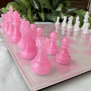 May include: A pink and white chess set with glitter on the pieces. The board is made of clear plastic with a white and pink checkerboard pattern.