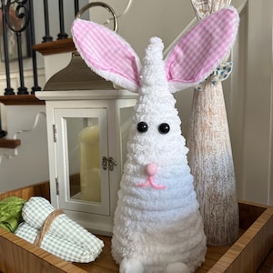 Puede incluir: Dos decoraciones de conejo blanco, una con orejas de vichy rosa y un cuerpo blanco esponjoso, la otra con un cuerpo de madera y orejas de vichy rosa. Los conejos están sentados en una bandeja de madera con zanahorias de vichy verde y blanco.