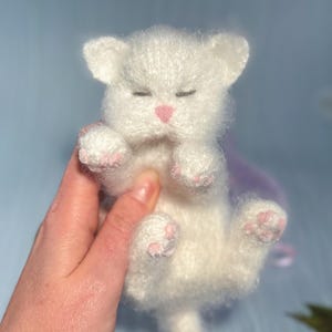 May include: A handmade white knitted cat toy with closed eyes and pink nose and paw pads. The cat is lying on its back, held by a hand. The background is a soft blue with hints of purple.