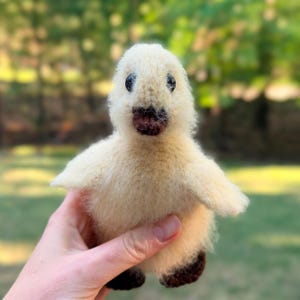 May include: A small, fuzzy, cream-colored needle-felted duckling figurine with a soft, textured appearance. The duckling features two small, dark, round eyes and a dark brown beak. The feet are also dark brown, contrasting with the light body.