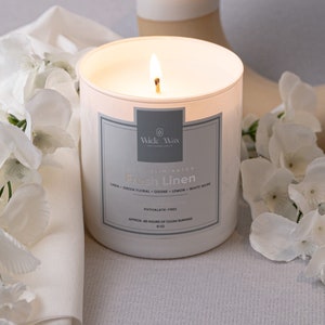 May include: A lit white candle with a label that reads "Fresh Linen" sits next to a white vase and white flowers. The candle is in a glass container. The vase is white and beige. The flowers are white and arranged around the candle and a white cloth.