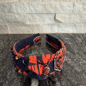 May include: A navy blue and orange fabric headband with a knot design. The headband is adorned with small blue beads and a decorative pendant. The fabric features a repeating pattern of orange shapes on a navy background.