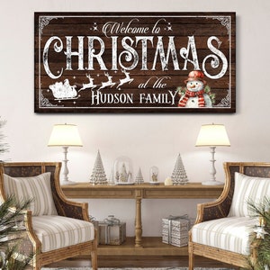 May include: A rustic wooden sign with white text that reads "Welcome to Christmas at the Hudson Family". The sign features a snowman with a red scarf and a sleigh with reindeer.
