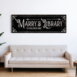 Può includere: Un cartello bianco e nero con il testo "Marry's Library Established 2022" su uno sfondo in legno invecchiato. Il cartello ha un bordo decorativo con accenti floreali.