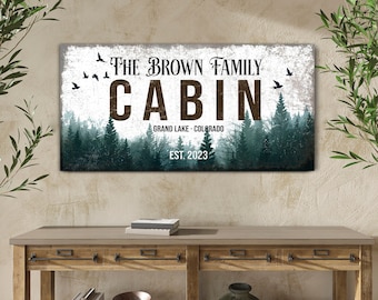 May include: A rustic wooden sign with a white background and distressed brown text. The sign reads "The Brown Family Cabin Grand Lake - Colorado Est. 2023". The sign is surrounded by a green forest background with birds flying in the sky.