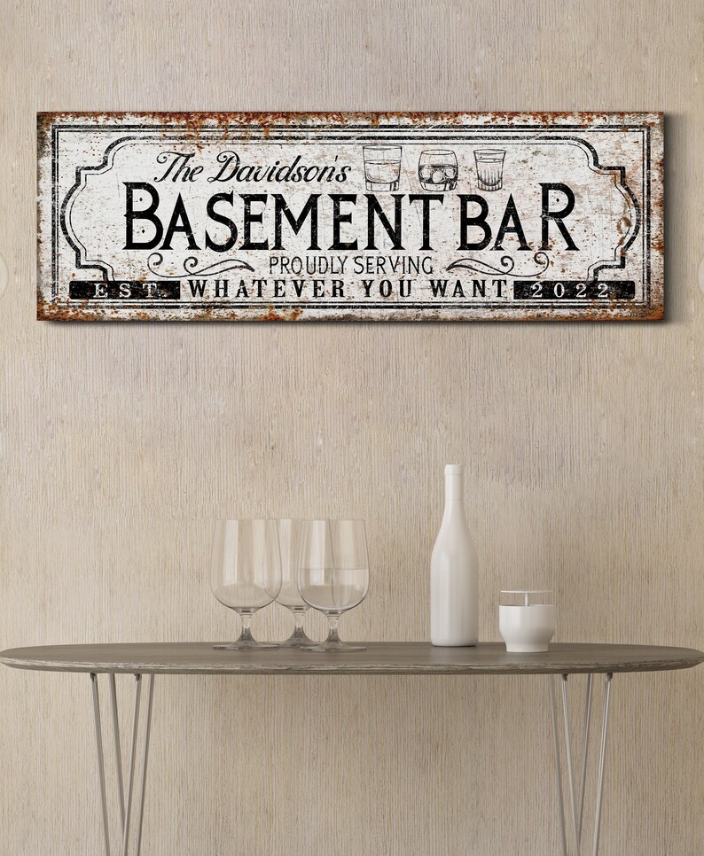 May include: A white sign with black text that reads "The Davidsons Basement Bar Proudly Serving Whatever You Want Est. 2022". The sign has a rustic, distressed look and features three glasses of alcohol.