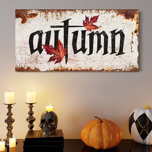 Op de afbeelding: Een rustiek houten bord met het woord "autumn" in zwarte gotische letters, versierd met rode esdoornbladeren. Het bord staat boven een tafel met kaarsen, een schedel en pompoenen, wat een herfstthema cre&euml;ert.