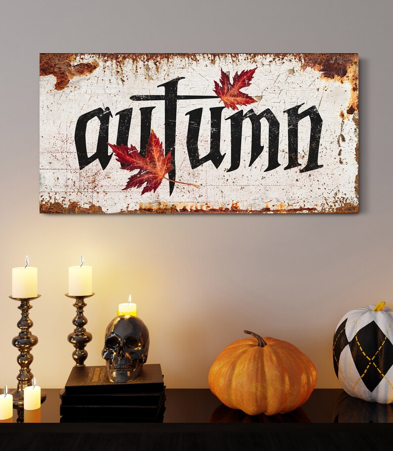 Op de afbeelding: Een rustiek houten bord met het woord "autumn" in zwarte gotische letters, versierd met rode esdoornbladeren. Het bord staat boven een tafel met kaarsen, een schedel en pompoenen, wat een herfstthema cre&euml;ert.