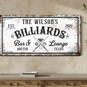 May include: A white sign with black text that reads "The Wilson's Billiards Bar & Lounge Est. 2022 Austin Texas". The sign has a distressed look and features a crossed pool cue and billiard balls.
