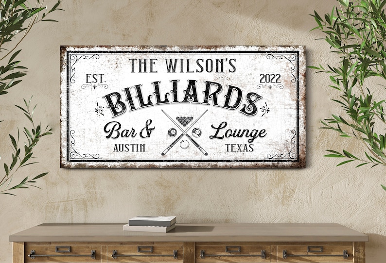 May include: A white sign with black text that reads "The Wilson's Billiards Bar & Lounge Est. 2022 Austin Texas". The sign has a distressed look and features a crossed pool cue and billiard balls.