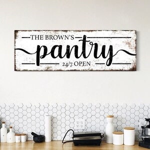 May include: A white wooden sign with black text that reads "The Brown's Pantry 24/7 Open". The sign has a distressed look and is hanging on a white wall above a kitchen counter.