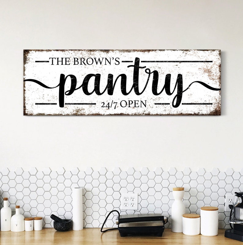 May include: A white wooden sign with black text that reads "The Brown's Pantry 24/7 Open". The sign has a distressed look and is hanging on a white wall above a kitchen counter.