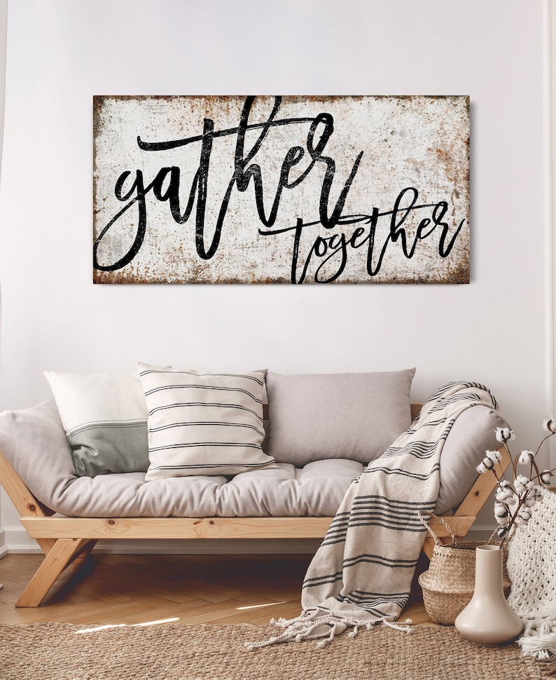 May include: A white wooden sign with black lettering that reads "gather together". The sign has a distressed, rustic look and is hanging on a white wall above a light gray couch with pillows and a throw blanket.