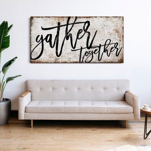 May include: A white wooden sign with black lettering that reads "gather together". The sign has a distressed, rustic look and is hanging on a white wall above a light gray couch.