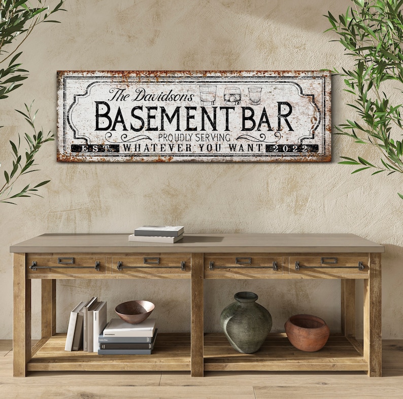 May include: A white sign with a rusty brown border and black text that reads "The Davidsons Basement Bar Proudly Serving Whatever You Want Est. 2022". The sign has a black and white illustration of three glasses of alcohol.