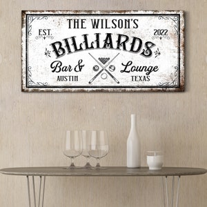 May include: A white sign with black text and graphics that reads "The Wilson's Billiards Bar & Lounge Austin Texas Est. 2022". The sign has a distressed, vintage look and features crossed pool cues and billiard balls.