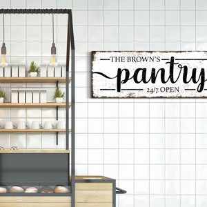 May include: A white wooden sign with black text that reads "The Brown's Pantry 24/7 Open". The sign is hanging on a white tiled wall in a kitchen.