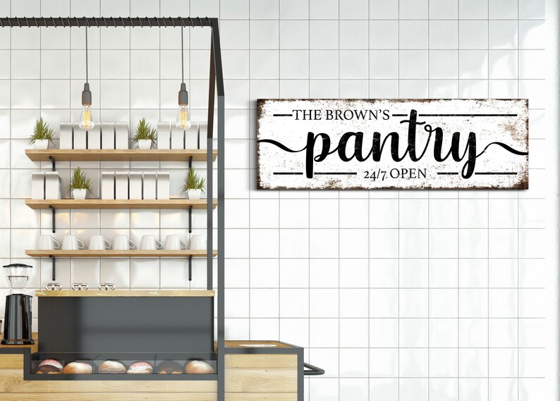 May include: A white wooden sign with black text that reads "The Brown's Pantry 24/7 Open". The sign is hanging on a white tiled wall in a kitchen.