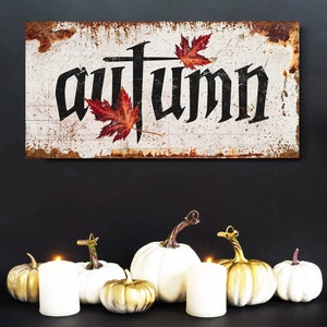 Op de afbeelding: Een rustiek bord met het woord "Autumn" in zwarte gotische letters, versierd met rode esdoornbladeren. Daaronder vormen witte en gouden pompoenen en brandende kaarsen een herfstthema.