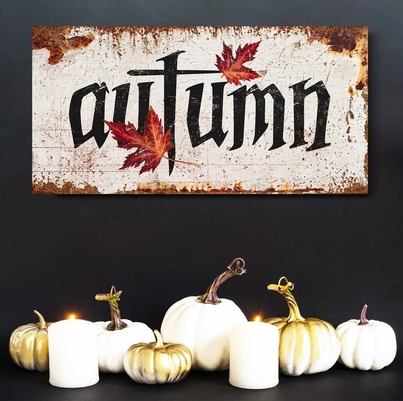 Op de afbeelding: Een rustiek bord met het woord "Autumn" in zwarte gotische letters, versierd met rode esdoornbladeren. Daaronder vormen witte en gouden pompoenen en brandende kaarsen een herfstthema.