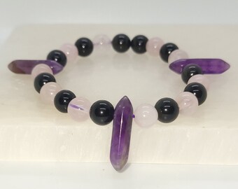 May include: A bracelet made with black, pink, and purple gemstone beads. The bracelet has two purple amethyst points hanging from it.
