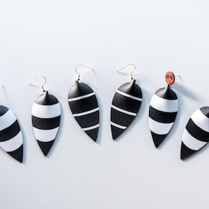 May include: Six black and white striped earrings with silver hooks. The earrings have a teardrop shape with a black top and white stripes. Two earrings have small red and black accents at the top. The earrings are displayed on a white surface.