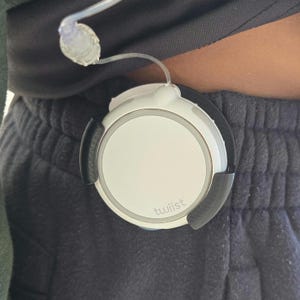 May include: A white and black medical device with the word "twiist" printed on it. The device is connected to a clear tube and is attached to a person's clothing. The device is circular with a white center and black accents.