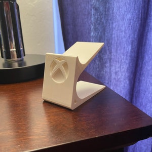 May include: White plastic stand with a cutout of the Xbox logo. The stand is designed to hold a video game console.