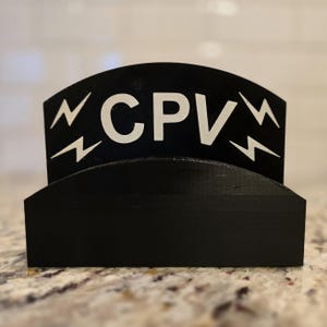 May include: A black, arched desk organizer with the letters "CPV" in white, flanked by white lightning bolt designs. The organizer sits on a speckled countertop.