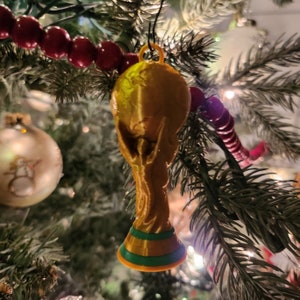 May include: A gold-colored trophy ornament with green and gold accents hanging on a Christmas tree.
