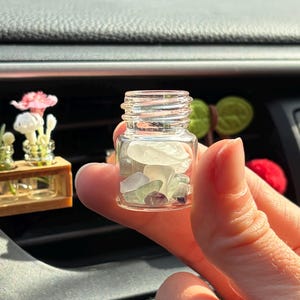Könnte beinhalten: Ein kleines, klares Glasgefäß gefüllt mit Meeresglas und kleinen lila Steinen. Das Glas wird in einer Hand gehalten, mit einem verschwommenen Hintergrund des Armaturenbretts eines Autos. Miniatur-Blumenarrangements sind im Hintergrund zu sehen.