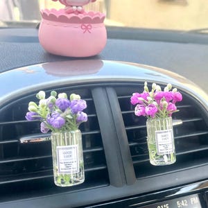 Puede incluir: Una figura de personaje de dibujos animados rosa se sitúa sobre dos clips para rejillas de ventilación de coche. Cada clip tiene un pequeño frasco de vidrio transparente con flores artificiales, una con flores moradas y otra rosas, con el texto GOOD LUCK.